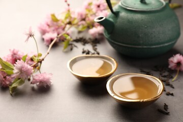 Obraz na płótnie Canvas Traditional ceremony. Cups of brewed tea, teapot and sakura tree branch on grey table, closeup
