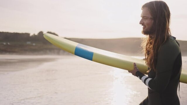 Young man carrying a surfboard walking on ocean beach. Surfer sports man getting ready to practice in the sea, catch a wave on a high tide. Active vacation evening or morning at the beach. 