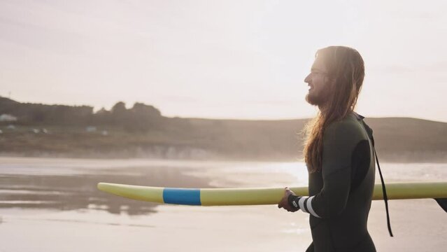 Young man carrying a surfboard walking on ocean beach. Surfer sports man getting ready to practice in the sea, catch a wave on a high tide. Active vacation evening or morning at the beach. 