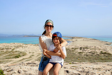 Portrait of mother holding her son with amazing Formentera's most iconic beach ("Ses illetes") on background, Formentera, Balearic Islands, Spain.