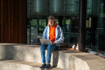 Student guy sitting on concrete bench against University building, studying online using laptop outdoors on sunny weather. Millennial man freelancer with computer outside. Freelance and urbanity