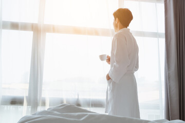 Tourist Asian man hold coffee cup in bathrobe towel looking at beautiful scene view on window at...