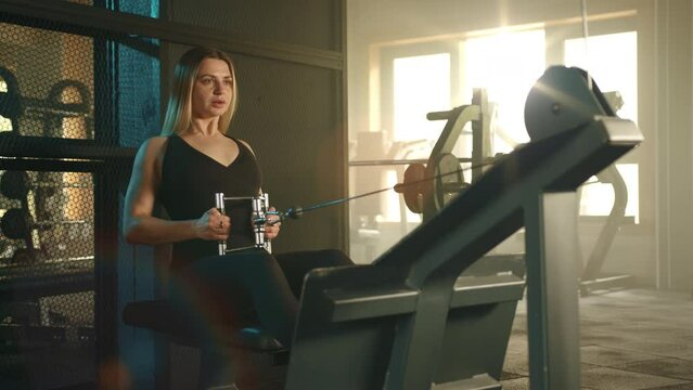 Pretty blonde with fit body in black sportswear performs seated cable row on cable machine. Woman exercises in modern fully-equipped gym lit by sunlight. High quality 4k footage