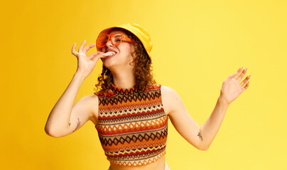 Portrait of pretty young girl in panama and knitted top eating candy floss against yellow studio background. Enjoyment. Concept of human emotions, youth culture, fashion, lifestyle, positivity