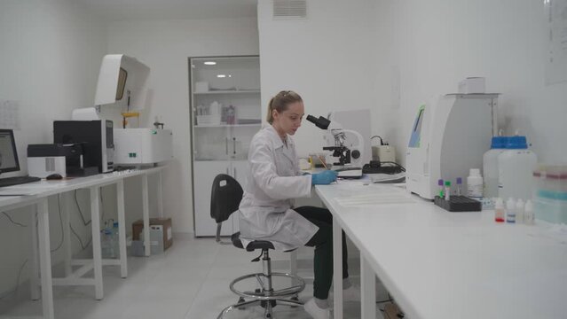 Female medic in uniform working with microscope making analysis at laboratory office. Scientist working in laboratory. Microbiologist woman working in modern medical lab with technological equipment.