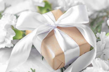 Fototapeta na wymiar Gift box wrapped in craft paper with white bow on flowers