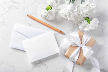 White paper card and envelope with gift box and flowers on marble background