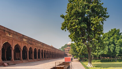 Fototapeta premium The ancient gallery is built around the perimeter of the Taj Mahal complex. Colonnade of red sandstone with arches. A tower with a dome in the distance. In the garden on the lawn - green grass, trees