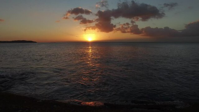drone flies low over rocks toward sea and sunrise.
