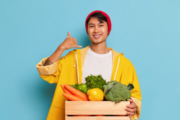 Smiling guy in red hat and yellow coat poses against blue background, box with vegetables in front...