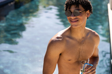 Happy shirtless biracial fit man in sunglasses drinking cocktail standing in swimming pool in sun