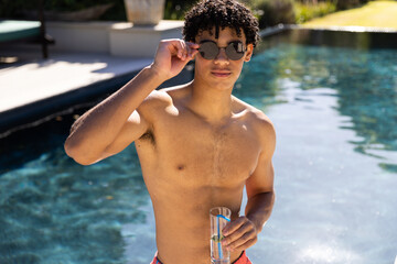 Portrait of shirtless biracial fit man in sunglasses drinking cocktail by in swimming pool in sun