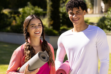 Portrait of happy fit diverse couple standing in sunny garden holding yoga mats