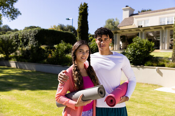 Portrait of happy fit diverse couple standing in sunny garden embracing and holding yoga mats