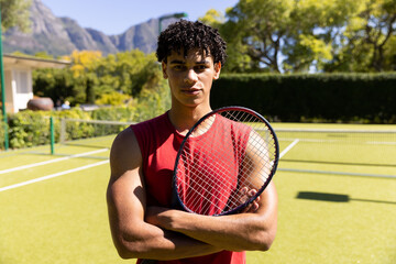 Portrait of happy biracial fit man holding tennis racket on sunny outdoor tennis court
