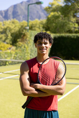 Portrait of happy biracial fit man holding tennis racket on sunny outdoor tennis court