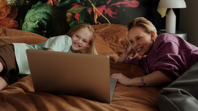 Modern Caucasian woman and her preteen daughter relaxing together on bed at home enjoying watching funny videos in Internet on laptop