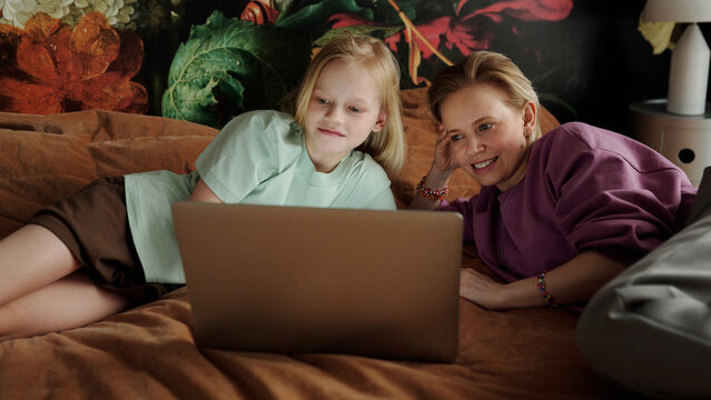 Caucasian woman and her preteen daughter relaxing together on bed at home watching series online on laptop