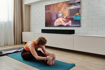 Modern Caucasian woman with short hair practicing yoga at home doing seated forward bend asana with online fitness trainer on TV screen