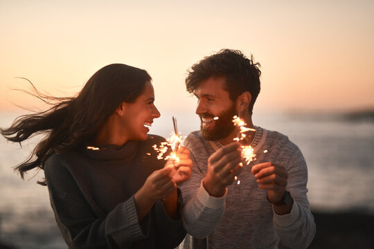 Love, beach and couple with sparklers for celebration, party and quality time on romantic date. Nature, sunset and happy man and woman with fireworks for honeymoon on holiday, weekend and vacation - Powered by Adobe