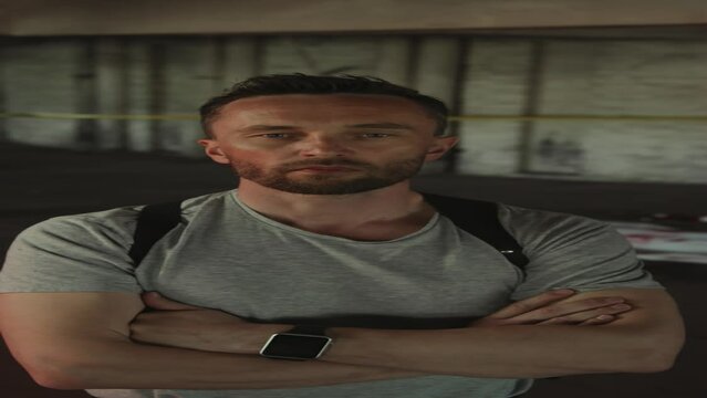 Medium vertical portrait shot of grim Caucasian male detective in plain t-shirt and gun harness standing with crossed arms and posing at murder scene, flashing police lights and corpse in background
