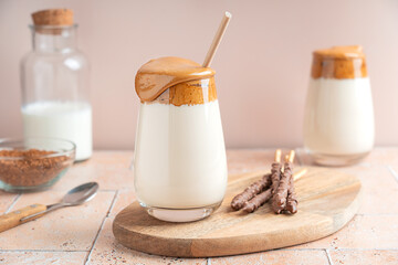 Homemade sweet dalgona cold drink made of whipped instant coffee, sugar and water topped on milk served in glass with paper straw and chocolate sticks on wooden board on stony table with ingredients