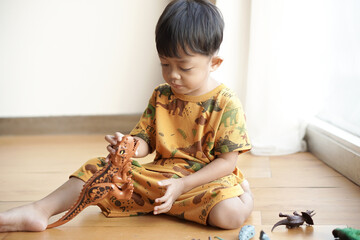 Handsome Asian toddler happily playing with a plastic dinosaur toy near the window in a hotel