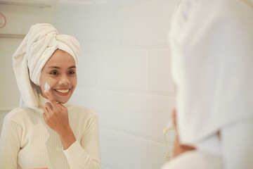 Beautiful young asian woman in white robe applying lotion on face in bathroom in front of big mirror