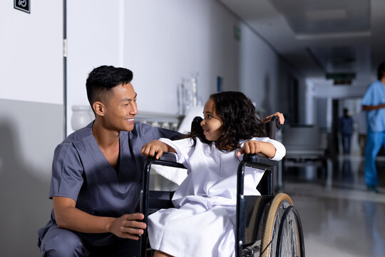 Smiling asian male doctor and biracial girl patient in wheelchair talking in corridor at hospital