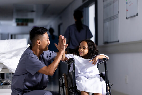 Happy asian male doctor and biracial girl patient in wheelchair high fiving in corridor at hospital