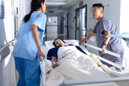 Diverse male and female doctor pushing female patient lying in bed in corridor at hospital