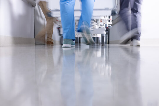 Low section of diverse doctors pushing patient in hospital bed down corridor, motion blur