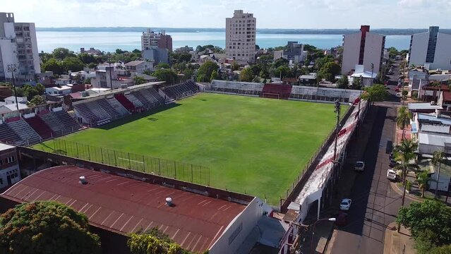 Aerial drone forward moving shot flying low over an empty football field of Club Deportivo Guaraní Antonio Franco in Argentina with the view of tall buildings in the background.