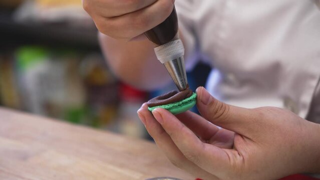 Pastry chef perfectly pipes chocolate cream filling onto French macaron cookie shell, slow motion close up 4K