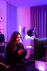Fotobehang Gaming Asian pretty gamer girl use joystick when playing video game on computer screen while sitting ergonomic chair in entertainment pink neon room  © MergeIdea