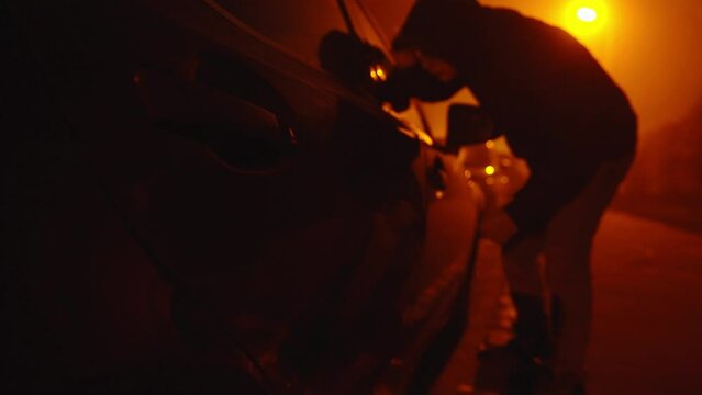 Hooded man trying to break into a car under orange street light, locked off