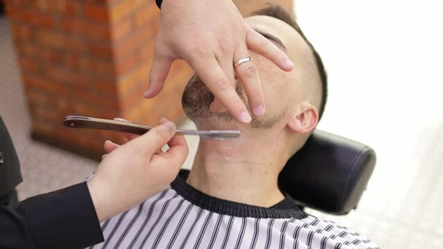 Barber Man is shaving client with a vintage straight razor in a barbershop. Classic shave by stainless steel straight edge razor