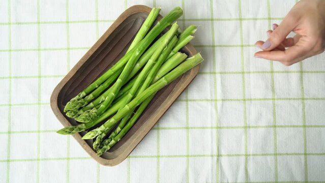 Woman hands puts a wooden bowl with fresh asparagus on a checkered tablecloth. Bunches of vegetable in basket top view close up. Spring season, new harvest of dutch or german green asparagus. 4k 25FPS