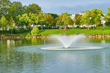 landscape city park. City pond in the park with a geyser fountain in the water. Coastline with green grass and trees