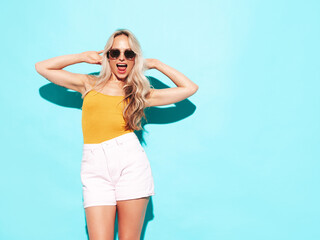 Young beautiful smiling blond female in trendy summer shorts clothes. Sexy carefree woman posing near blue wall in studio. Positive model having fun. Cheerful and happy. Going crazy