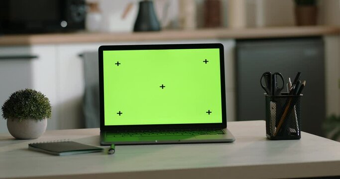 A laptop on table with green screen on background of white light kitchen set. Template for inserting any video with laptop computer on the desk at home. Mockup, a laptop template with a green screen.