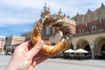 Hand holding obwarzanek krakowski pretzel on Kraków Main Market Square, with Cracow Cloth Hall...