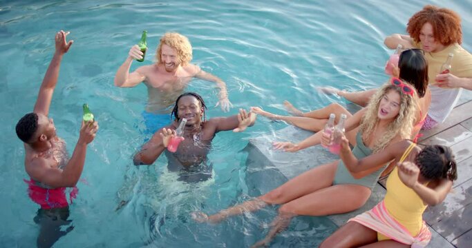 Happy diverse friends with drinks dancing and swimming in pool in slow motion