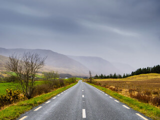 Small narrow asphalt road to mountains in low cloudy sky. Travel background. Calm Irish nature landscape. Transportation and tourism concept. West of Ireland.