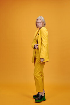 Vertical full body studio portrait of stylish mature woman with blond hair wearing elegant suit and mules and posing on camera, bright yellow background