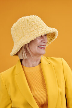 Vertical medium close-up portrait of modern mature Caucasian woman wearing stylish yellow outfit with hat looking away, bright yellow background