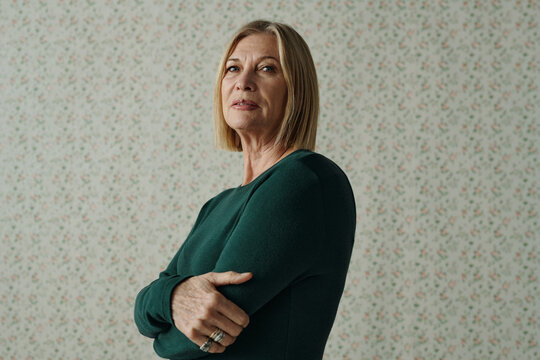 Horizontal medium portrait of beautiful senior Caucasian woman wearing dark tidewater green sweater standing against flower pattern wallpaper background