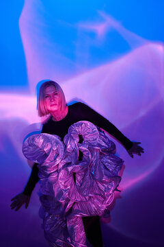 Vertical studio shot of stylish senior Caucasian woman wearing futuristic clothes posing on camera in neon light, generation Z style concept
