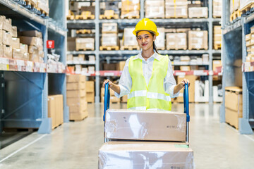 Asian woman warehouse worker in uniform walking through a warehouse while pushing trolley with cardboard boxes ready for delivery, cute girl employee working in stockroom with handcart