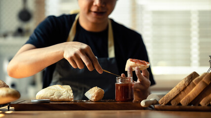 Man preparing a healthy breakfast, spreading raspberry jam on slice of bread with table knife at kitchen table
